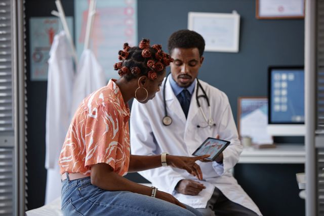 Schwarze Frau zeigt im Gespräch mit dem Arzt auf den Bildschirm und stellt Fragen