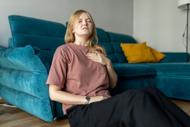 Frau sitzt mit starken Brustschmerzen und einer Hand, die ihre Brust berührt, während sie zu Hause Probleme hat. Herzinfarktkonzept oder Herzinsuffizienzsymptom. Druck in der Brust.