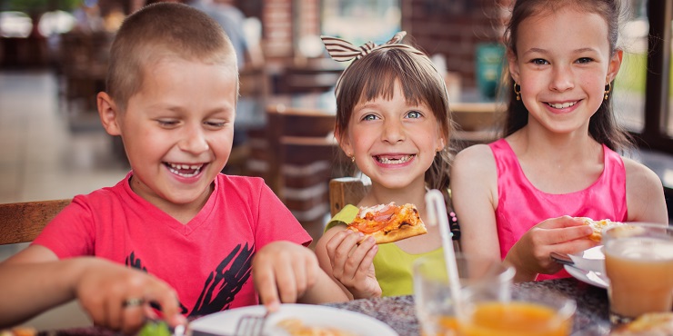 Symbolbild, drei Kinder zwischen 4 und 8 Jahre sitzen lachend nebeneinander am Tisch und essen Pizza.