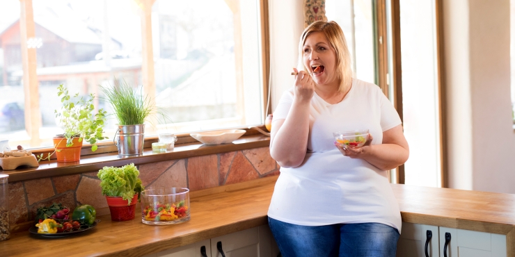 Symbolbild, übergewichtige Frau steht in der Küche und isst einen gesunden Salat