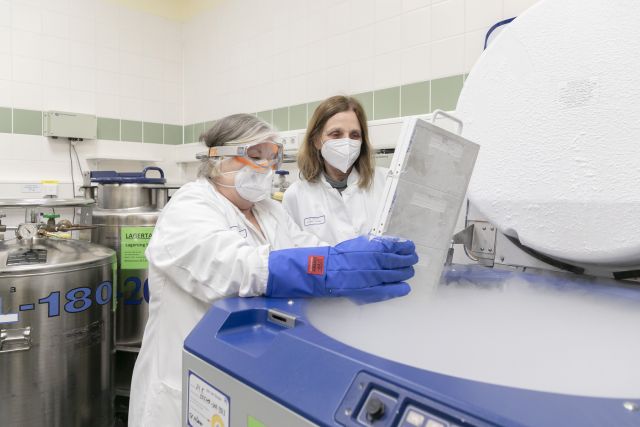 Das Bild zeigt zwei Forscherinnen im weißen Kittel, die an einem Laborgerät gekühlte Proben entnehmen.