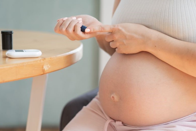 Schwangere Frau misst Blutzucker. Sichtbar: Babybauch, Hände, Glukosemessgerät auf Holztisch.