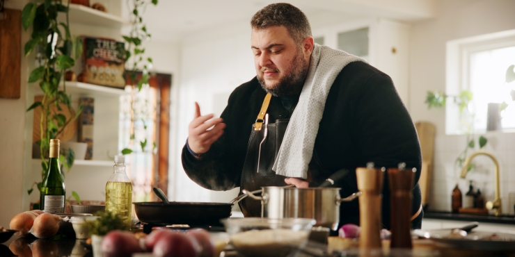 Symbolbild, adipöser Mann steht in der Küche und kocht sich ein Essen und riecht an den Dämpfen.