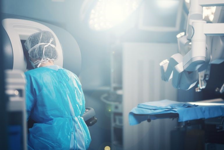 Chirurg in blauem OP-Kittel und Haube steuert Da-Vinci-Operationsroboter im hell beleuchteten OP-Saal.