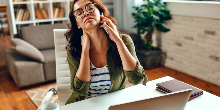 Symbolbild, Frau mit Nackenschmerzen sitzt am Schreibtisch vor ihrem Laptop und hält sich das Smartphone ans Ohr.