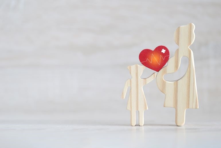 Holzfiguren von Schwangerer und Kind halten rotes Herz mit EKG-Linie auf hellem Hintergrund.