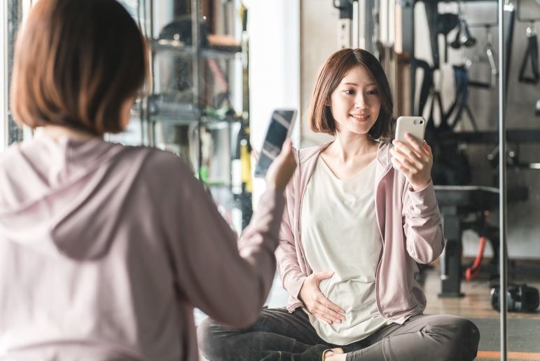 Schwangere Frau im Fitnessstudio recherchiert auf Smartphone. Rosa Jacke, Spiegelung zeigt Rückansicht.