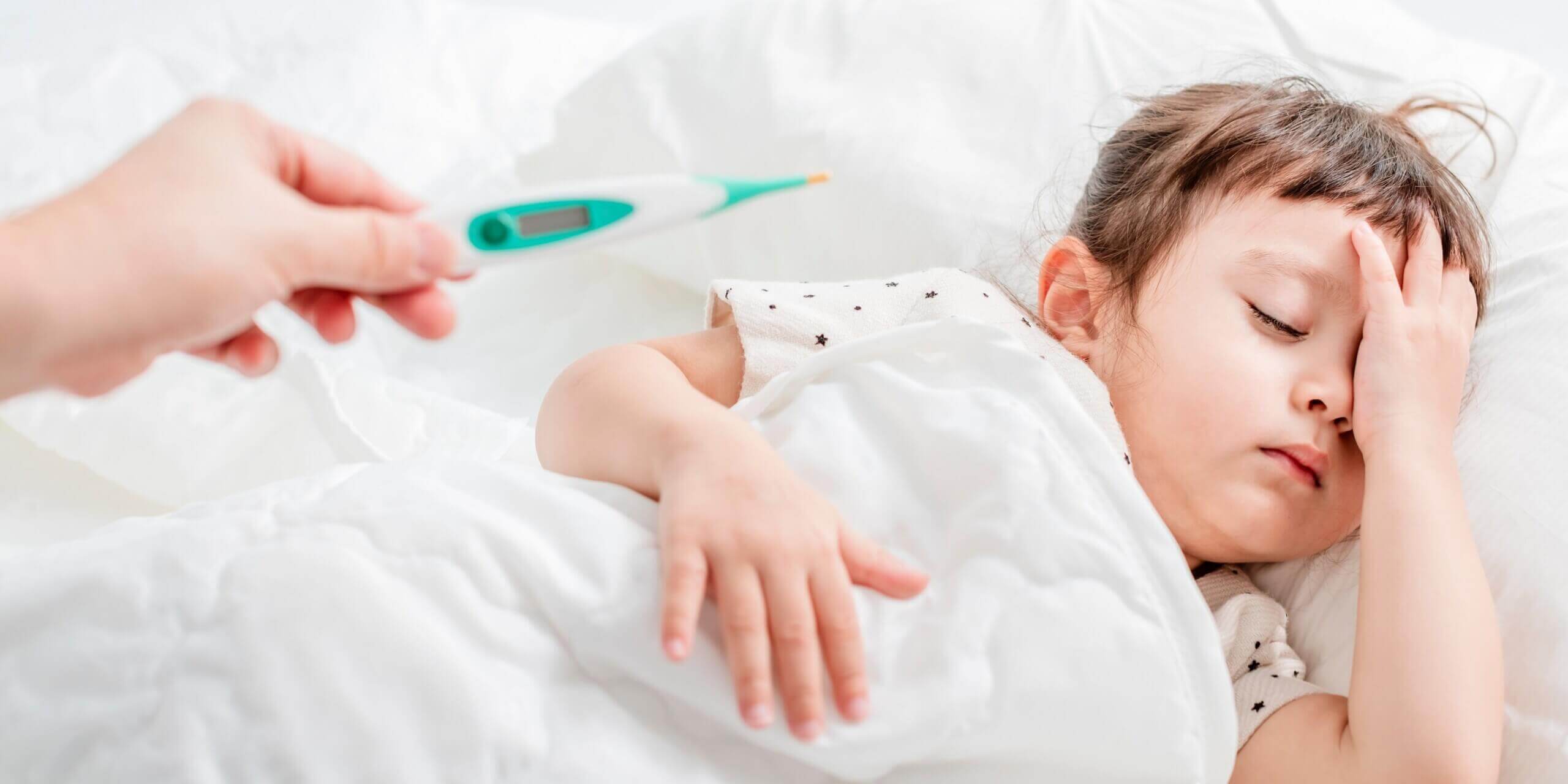 Kleinkind liegt mit Kopfschmerzen im Bett, Mutter hält Fieberthermometer