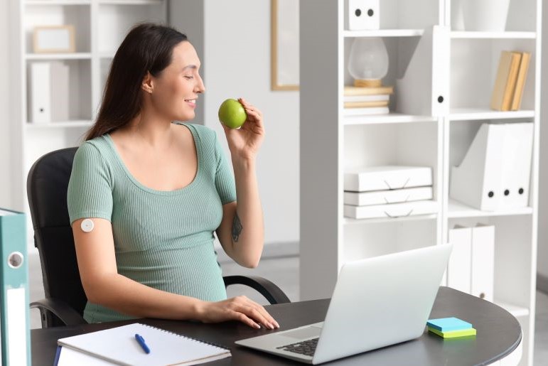 Schwangere Frau mit kontinuierlichem Glukosemesssensor für Gestationsdiabetes isst Apfel am Arbeitsplatz.
