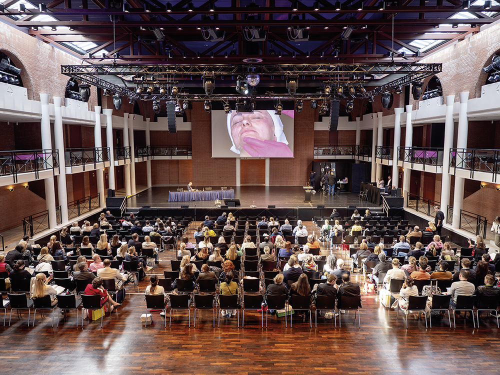 Ein großer Konferenzsaal mit Publikum, einer Bühne und einer Leinwand in einem modernen Veranstaltungsgebäude.