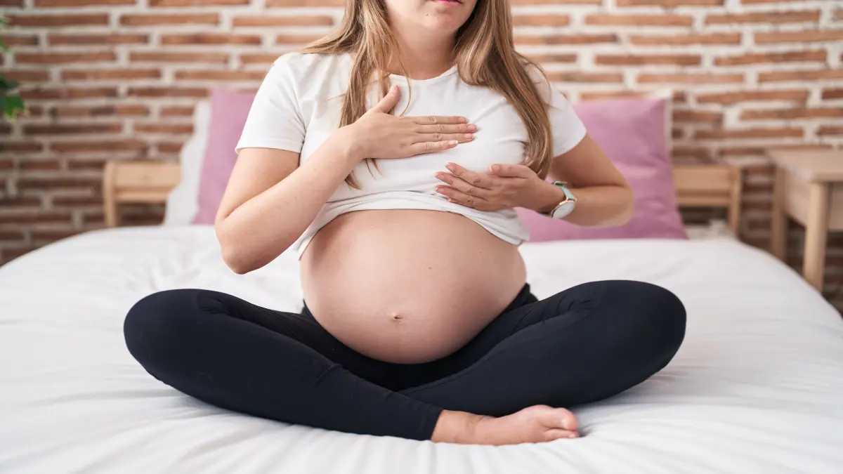 Junge schwangere Frau sitzt auf dem Bett und tastet ihre Brüste ab.