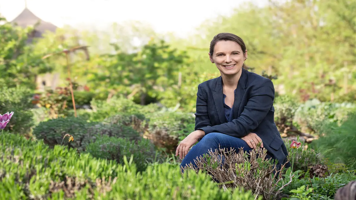Person sitzt in einem grünen Kräutergarten und trägt einen dunkelblauen Blazer.