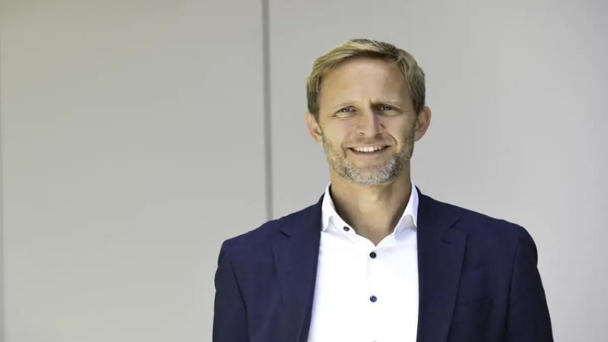 Person mit blonden Haaren in dunkelblauem Sakko und weißem Hemd vor hellem Hintergrund.