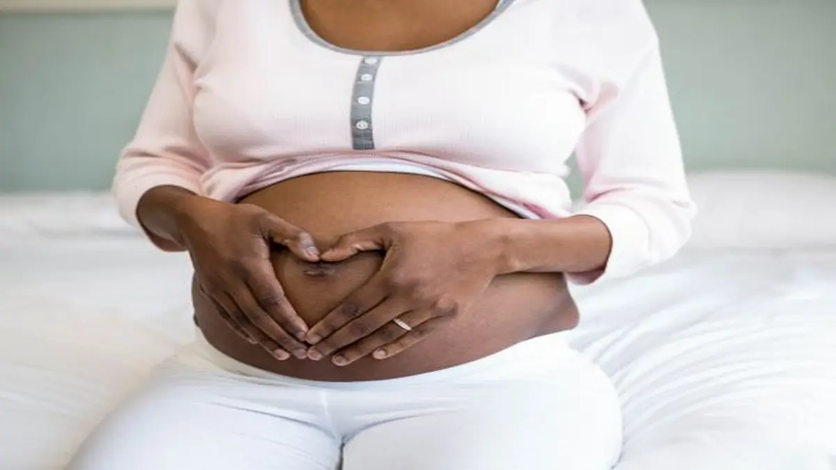 Schwangere Frau hält ihre Hände in Herzform über ihren Babybauch auf einem Bett.