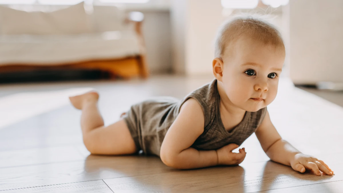 Baby in einem khakifarbenen Body krabbelt langsam über den Boden.