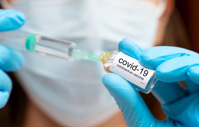 Nahaufnahme einer Hand in blauen Handschuhen, die eine Spritze und eine Ampulle mit der Aufschrift „COVID-19 Coronavirus Vaccine“ hält.