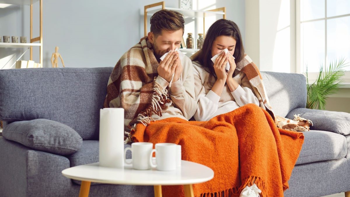 Ein Paar sitzt eingekuschelt in einer Decke auf dem Sofa, mit Tee und Taschentüchern, Symbol für Erkältung und Gemütlichkeit.