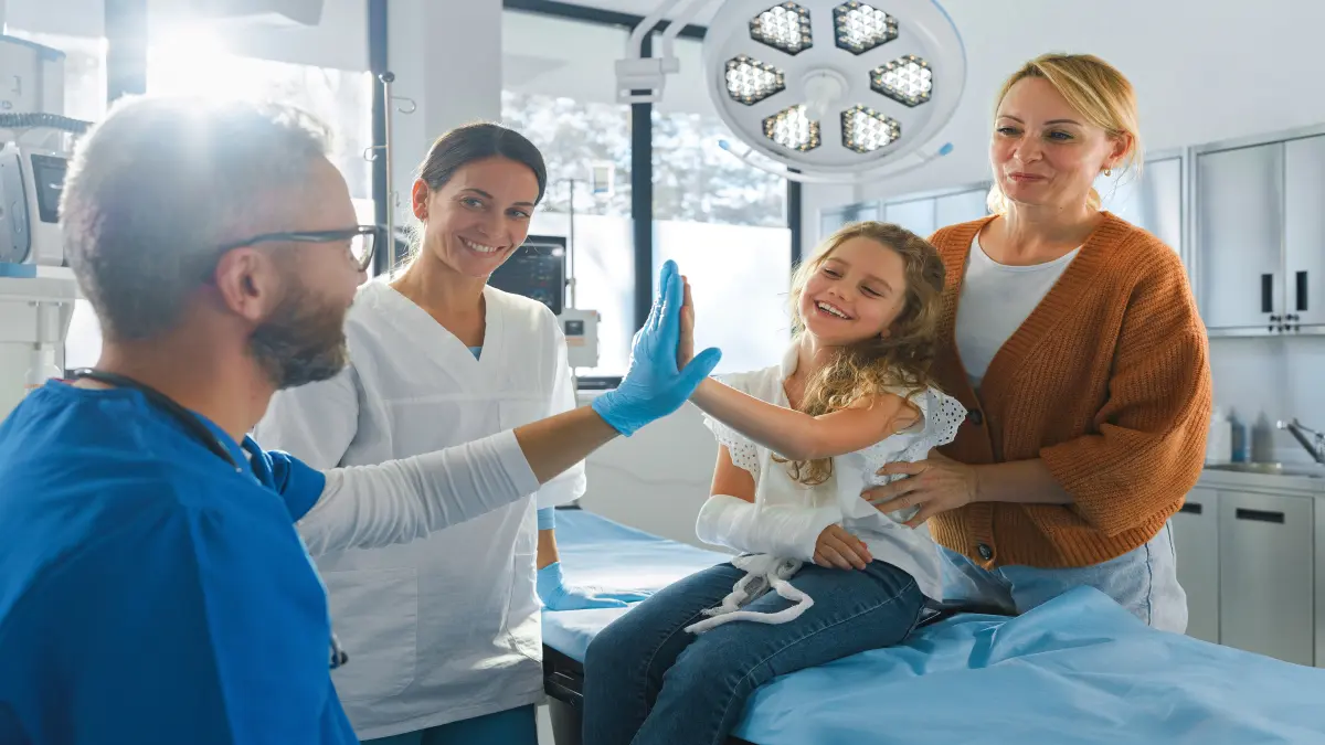 Mädchen und Arzt legen jeweils eine Hand aneindander und Mutter steht hinter dem Mädchen und eine Schwester daneben, sie befinden sich eine einem OP-Untersuchungsraum.