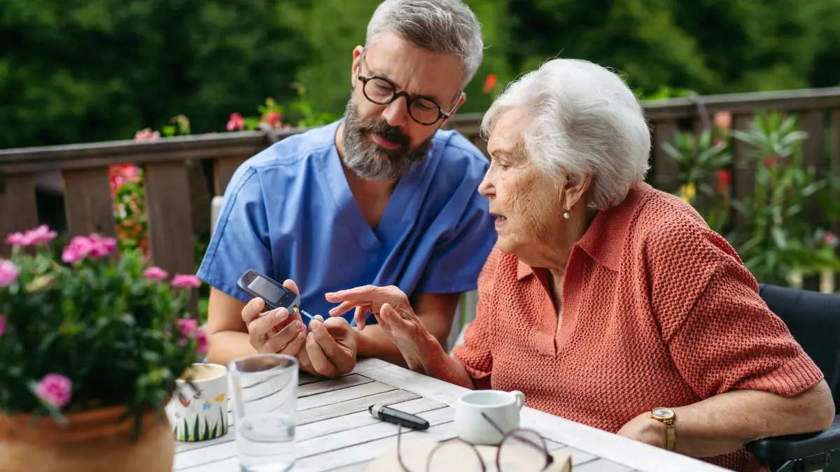 Ältere Dame mit Diabetes sitzt mit einem medizinischen Fachangestellten an einem Tisch und misst den Blutzuckerspiegel