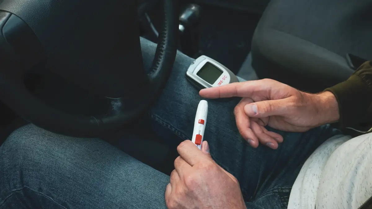 Ein Mann sitzt hinterm Steuer in einem Auto und misst mit einem Blutzuckermessgerät und einen Teststreifen in der Fingerkuppe seinen Blutzuckerspiegel.