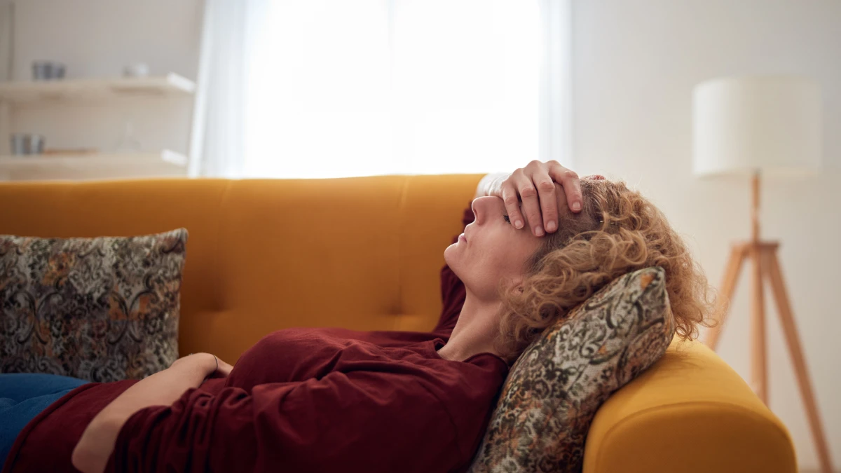 Frau liegt auf einem gelben Sofa und hält sich die Stirn. Symbolbild Kopfschmerz