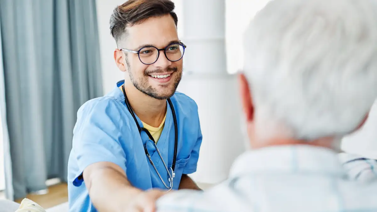 Ein lächelnder Pfleger trägt ein Stethoskop um den Hals und sitzt einem weißhaarigem Mann gegenüber und berührt ihn an der Schulter.