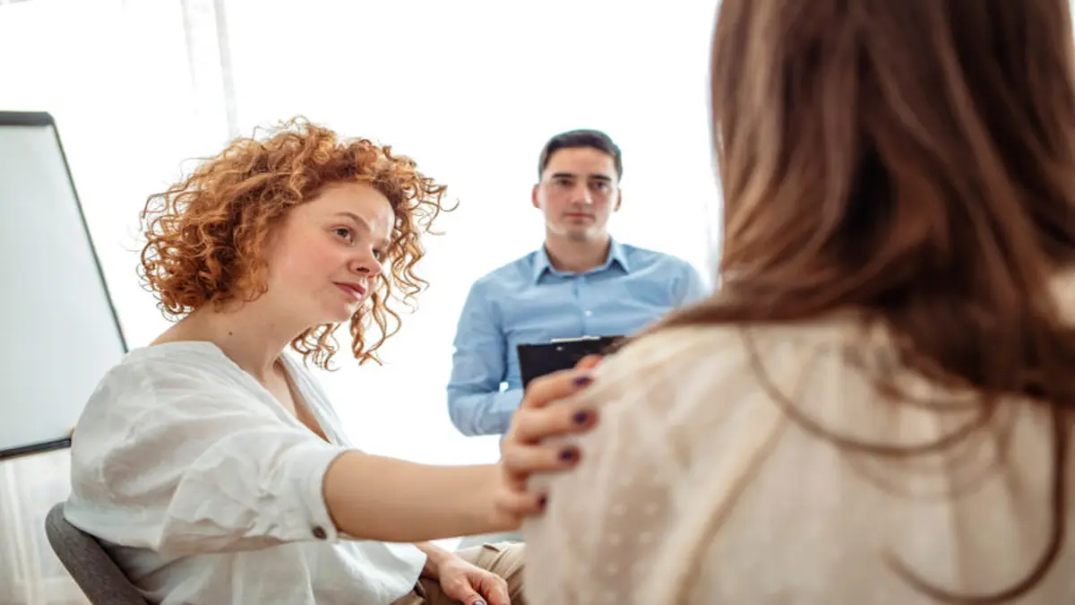 Zwei Frauen und ein Mann sitzen in einer Therapiesitzung, eine Frau legt unterstützend die Hand auf die Schulter der anderen