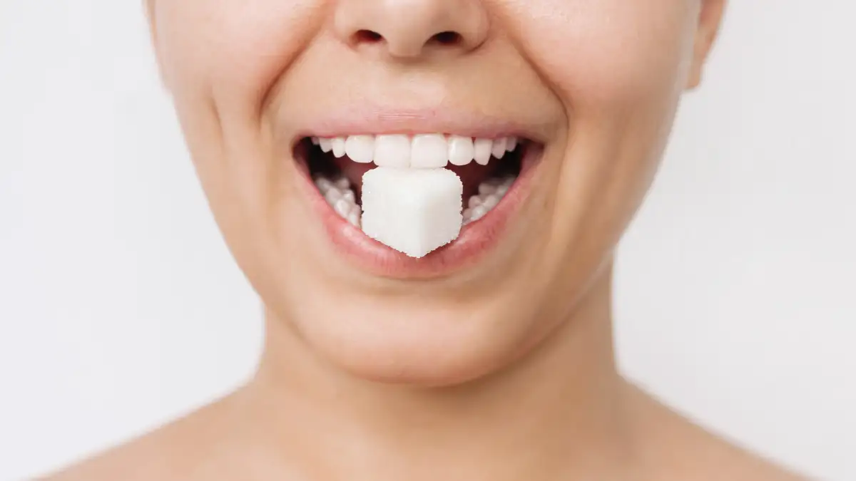 Das Gesicht einer Frau ist im Fokus, zwischen den weißen gesund Zähnen wird ein weißer Zuckerwürfel gehalten.