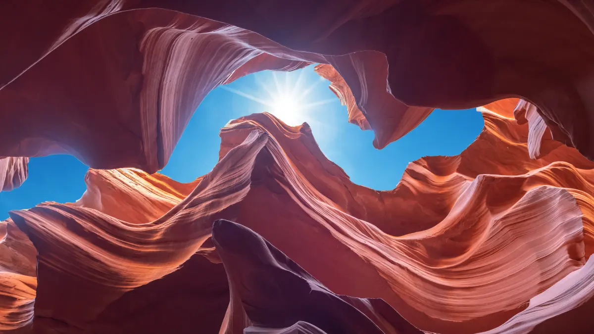 Blick aus einem Slot‑Canyon in Arizona hinauf zum blauen Himmel und Sonnenstern – geschwungene rote Sandsteinwände im Vordergrund