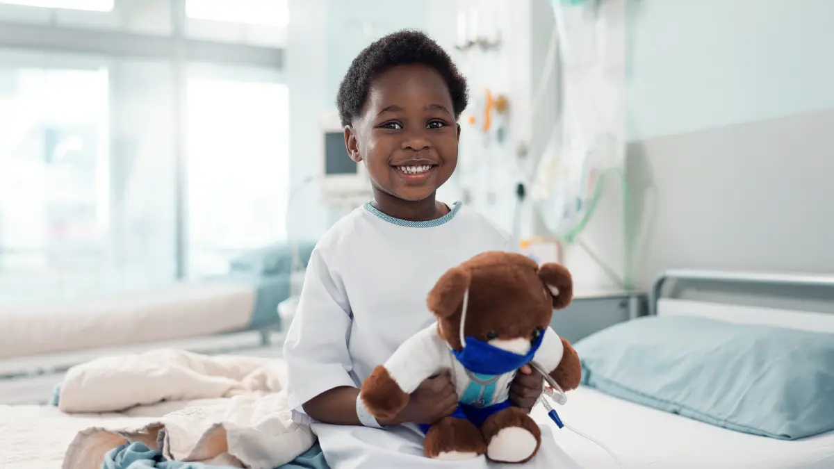 Kind im Krankenhausbett haelt einen Teddybear in medizinischer Kleidung