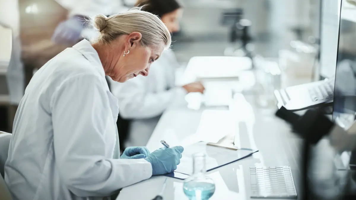 Wissenschaftlerin im weißen Kittel und blauen Latexhandschuhen notiert Daten auf einem Klemmbrett im Labor