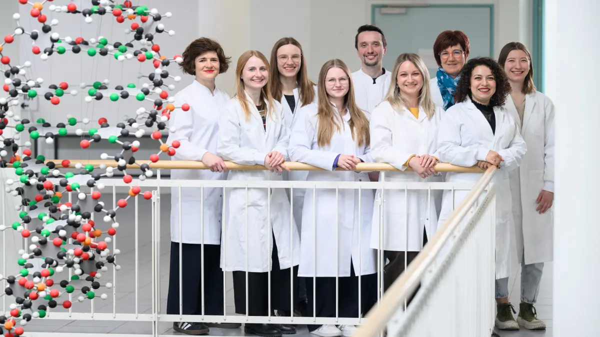 Forscherinnen und Forscher in Laborkitteln stehen als Arbeitsgruppe an einem Gelaender neben einem grossen DNA-Modell