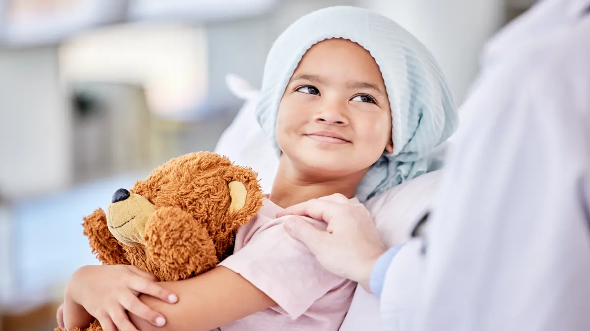Kind liegt im Krankenhausbett, haelt einen Teddybear im Arm und blickt laechelnd zu einer Person im Kittel