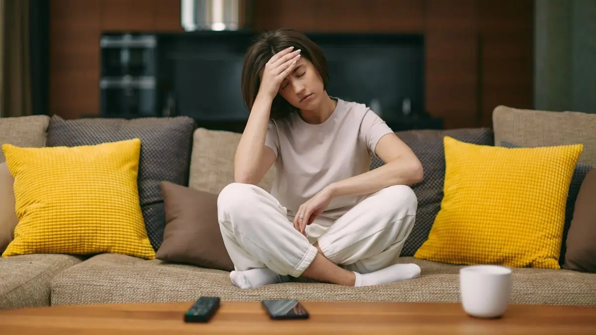 Erschoepfte Person sitzt auf dem Sofa und haelt sich die Stirn, Symbolbild fuer Kopfschmerzen nach Covid-19-Erkrankung und Impfung