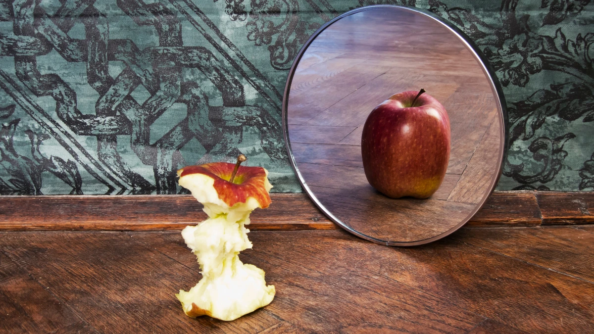 Abgebissener Apfelstrunk vor Spiegel, der einen ganzen Apfel zeigt – Symbol für Körperwahrnehmungsstörung bei Magersucht