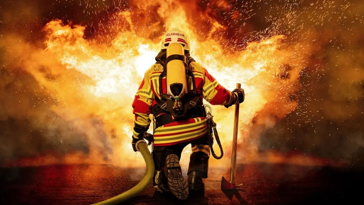Feuerwehrkraft kniet vor loderndem Feuer und bringt die Flammen unter Kontrolle, Symbolbild fuer PsO PsA und Systementzuendung im Griff