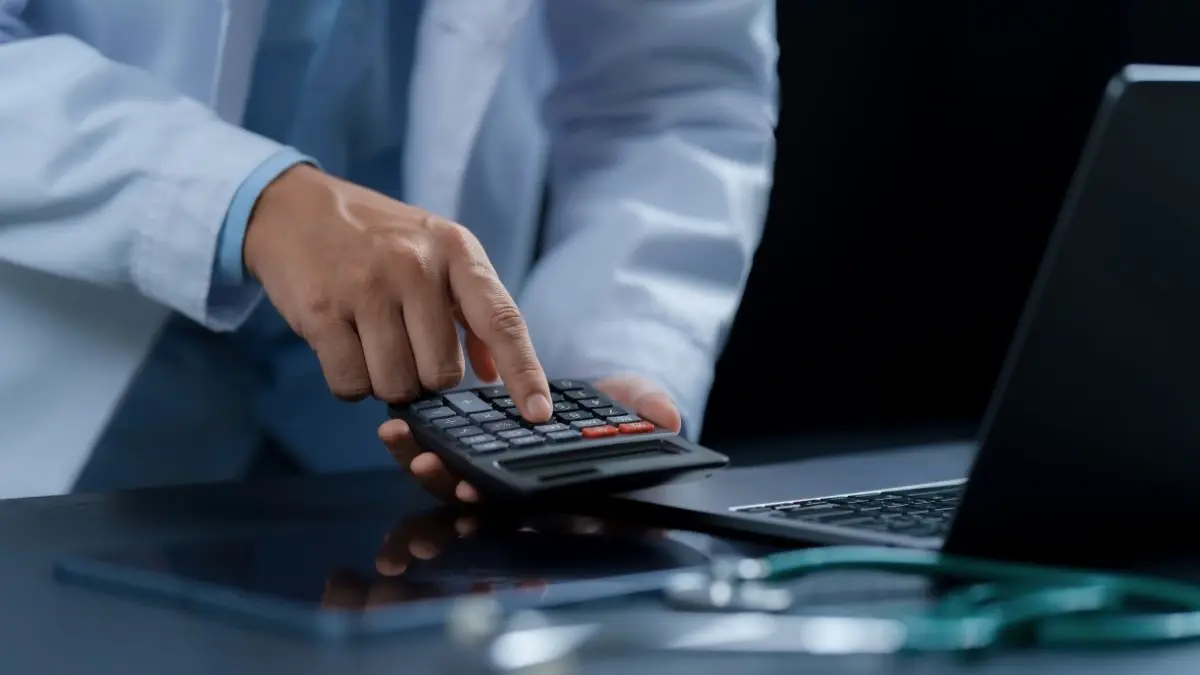 Person im Arztkittel arbeitet mit Taschenrechner neben Laptop und Stethoskop, Symbolbild fuer Rotstift Reform im Gesundheitswesen und gefaehrdete Patientenversorgung