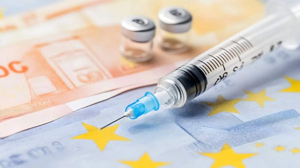 Spritze und Impfstoffflaschen vor EU-Flagge und Euro-Banknoten, Symbolbild fuer politische Debatte und Unmut ueber die Regierung