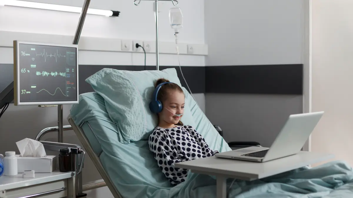 Mädchen liegt in Krankenhausbett, hat Kopfhörer auf und blickt auf einen Laptopbildschirm