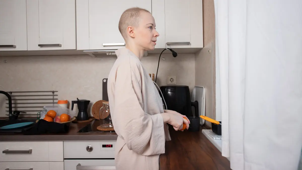 Eine kahlköpfige Frau in einem weichen Bademantel, die in ihrer Küche zu Hause sorgfältig eine Orange schält