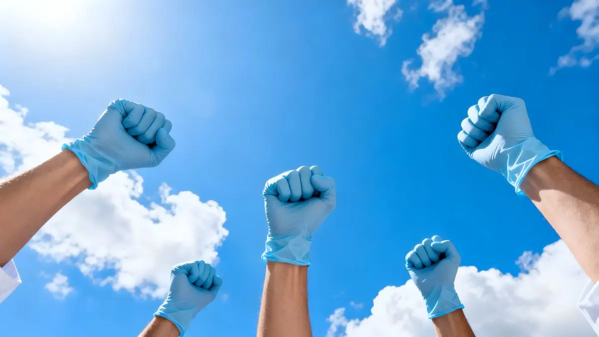 In die Hoehe gereckte Haende mit medizinischen Handschuhen vor blauem Himmel als Symbol fuer Forderungen des medizinischen Personals waehrend Gesundheitsminister ueber das GKV-Gesetz sprechen