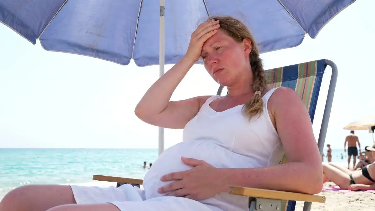 Schwangere Frau sitzt erschöpft unter Sonnenschirm am Strand als Symbol dafuer, dass Hitzeexposition das fetale Wachstum beeinflussen kann