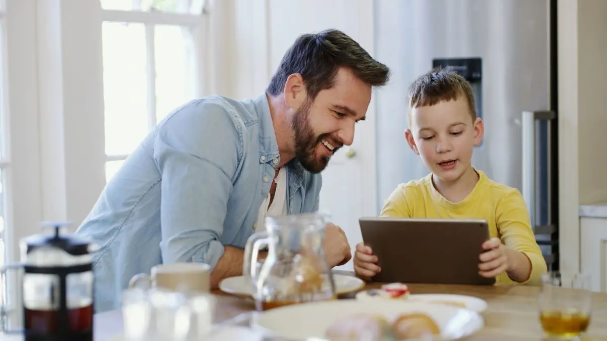 Vater erklaert seinem Sohn an einem Tablet spielerisch die Regeln der UV Praevention im Kindesalter