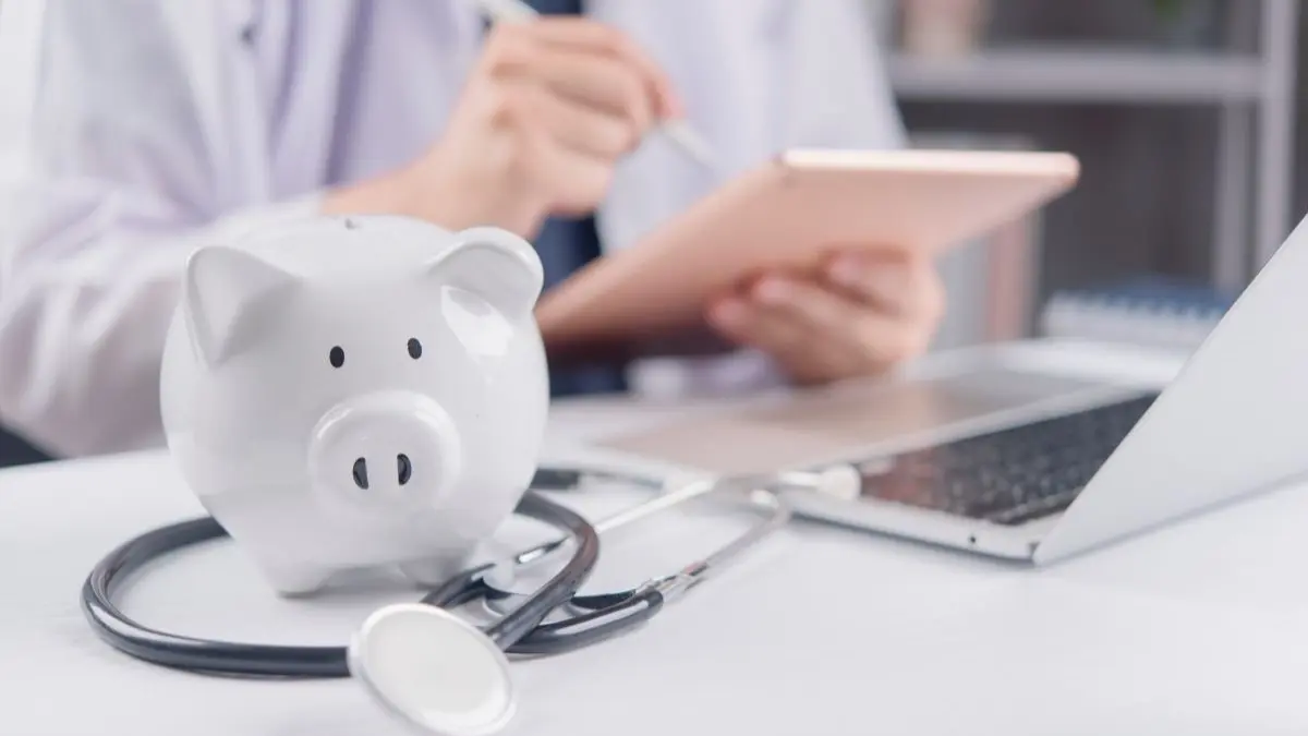 Weisses Sparschwein mit Stethoskop vor Laptop und Tablet als Symbol dafuer, dass das GKV Gesetz laut Rating Report viele Kliniken bis 2030 in die roten Zahlen treibt