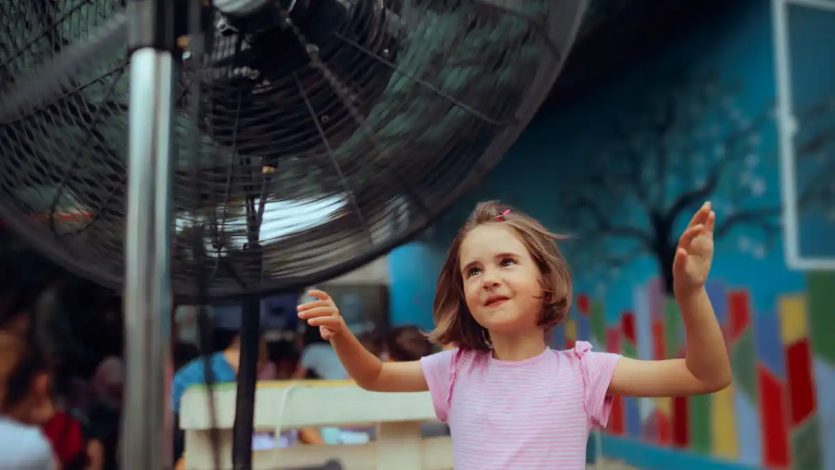 Auf dem Bild kühlt sich ein kleines Mädchen im Sommer vor einem elektrischen Ventilator, sie möchte damit wohltuende Erleichterung von der Sommerhitze finden.